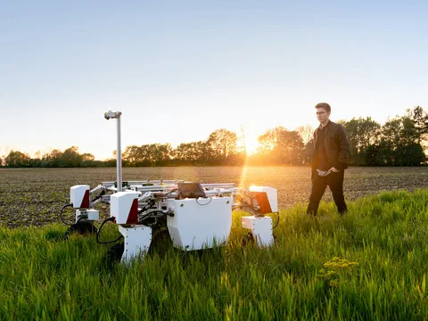 A field robot standing in tall grass at the edge of a crop field at sunset, a man walking beside it