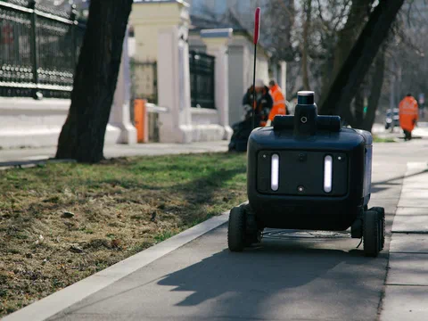 A six-wheeled delivery robot driving along a tree-lined city pavement